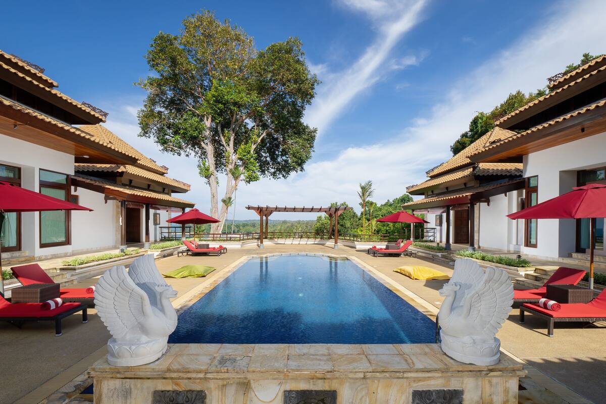 A swimming pool with two white statues, red lounge chairs, and umbrellas, surrounded by buildings with tiled roofs.