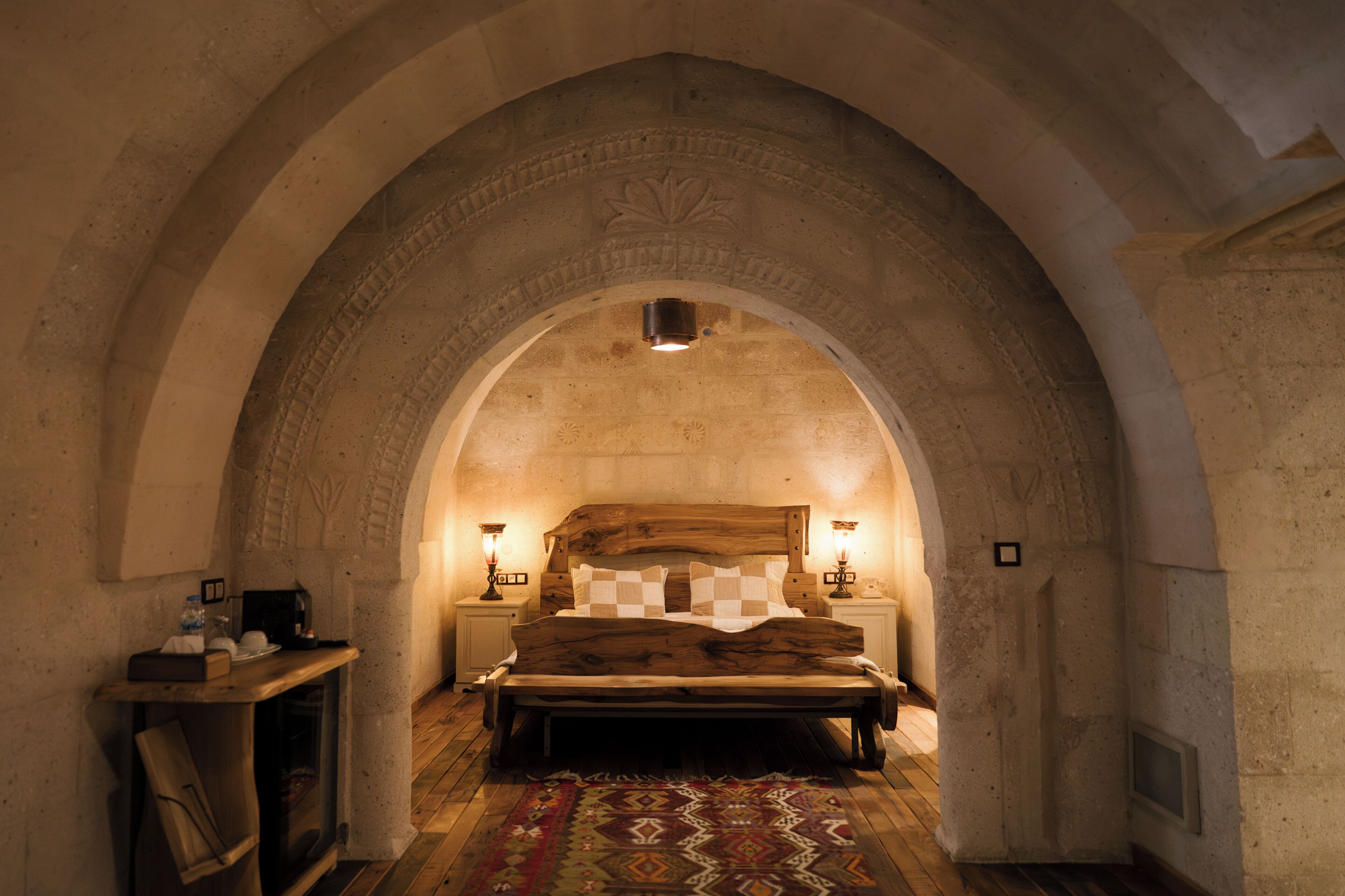 A room with a large bed under an arched stone ceiling, two bedside tables with lamps, and a wooden bench.