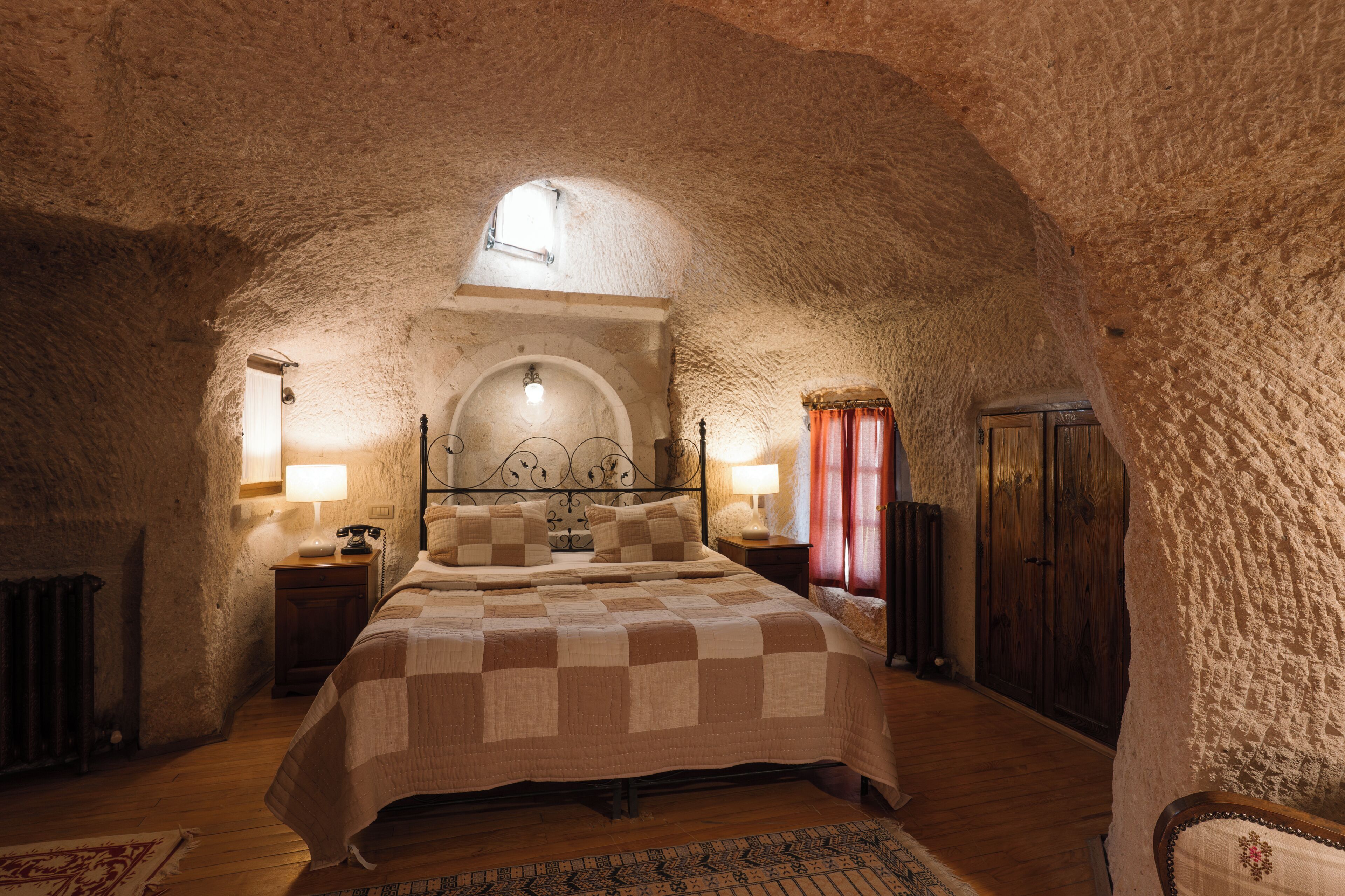 A bedroom with a checkered bedspread, a wooden floor, a stone wall, and a small arched window.