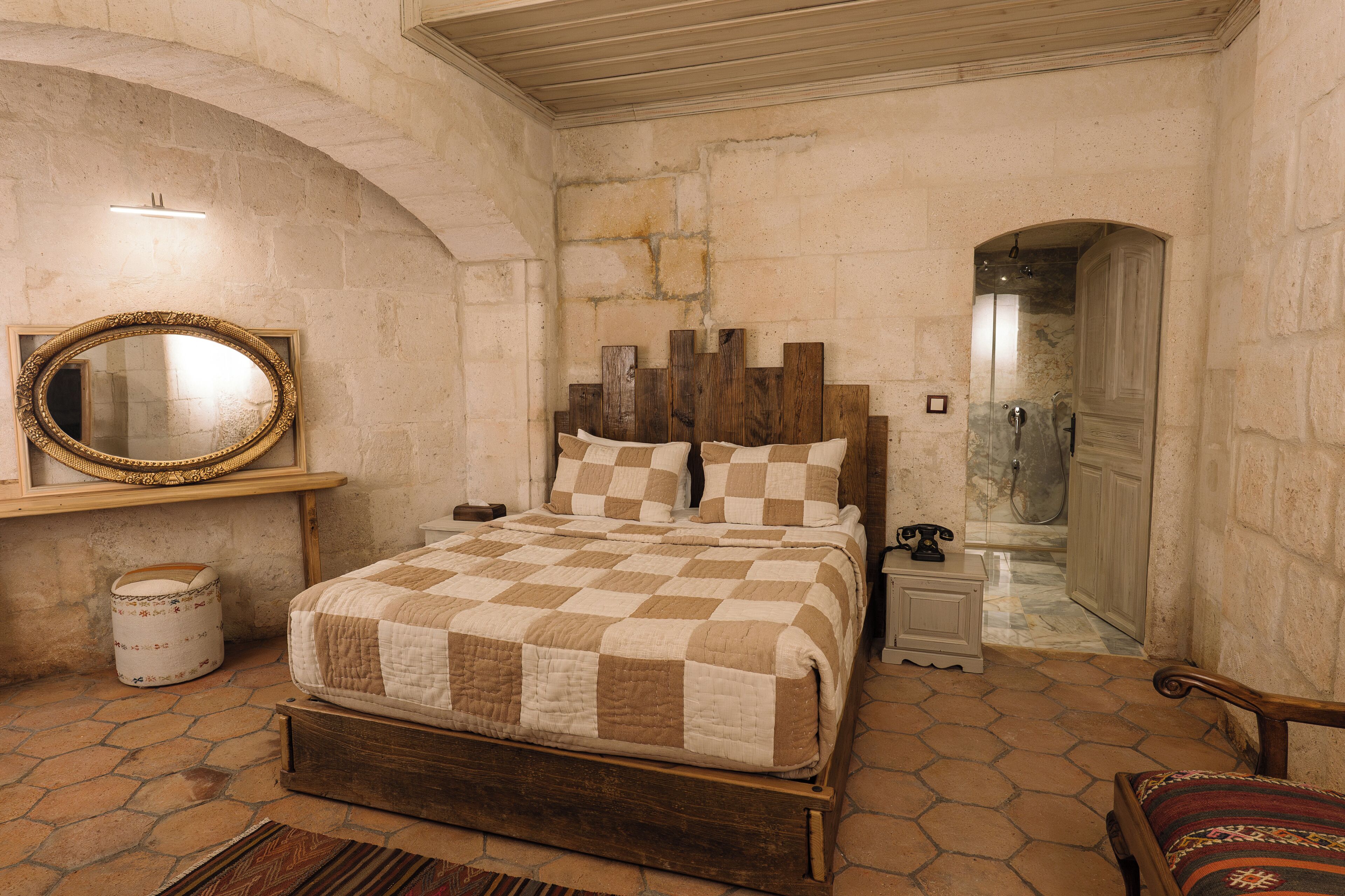 A bedroom with a checkered bedspread, a wooden headboard, a round mirror, and a stone wall.