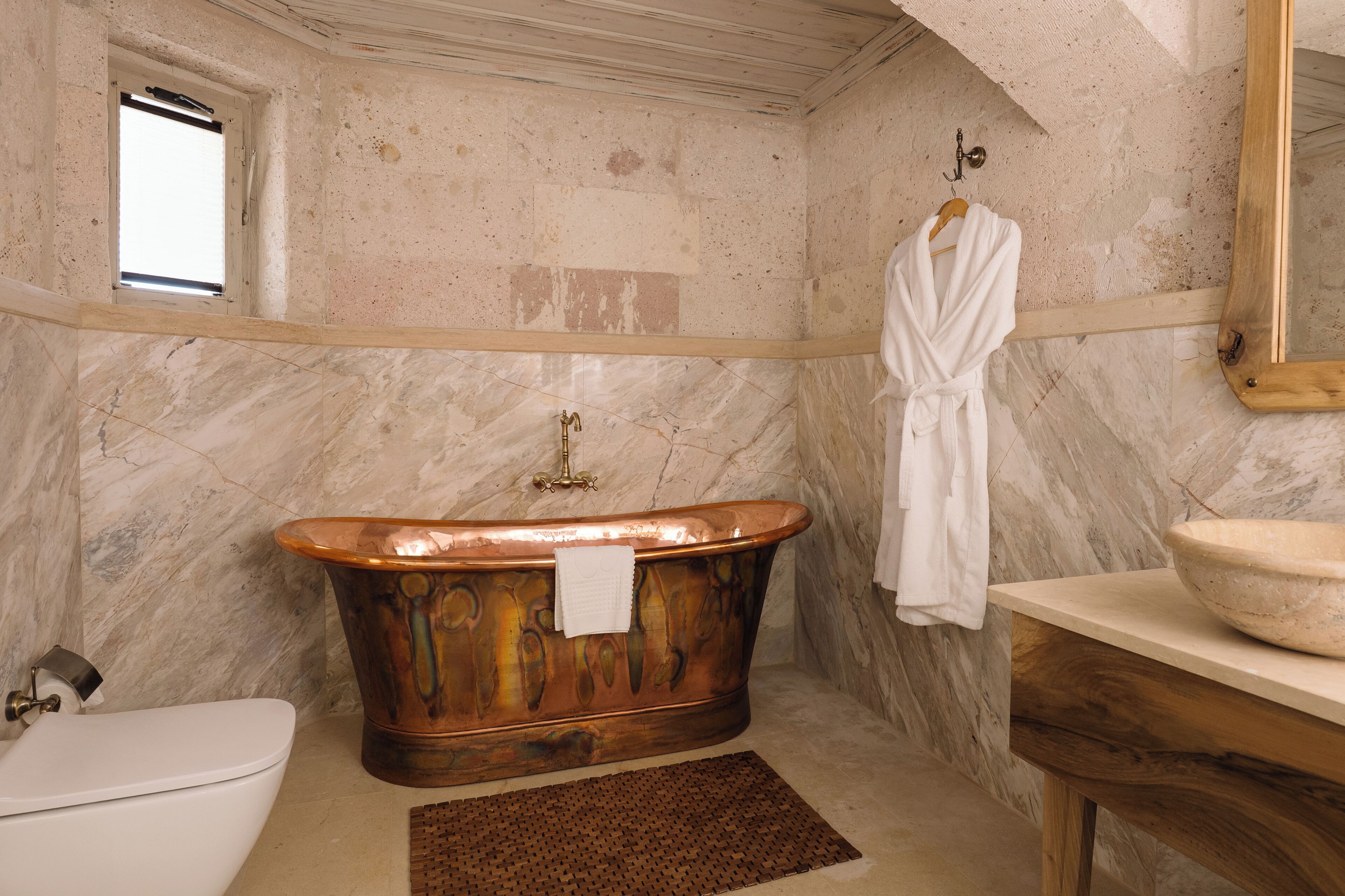 A bathroom with a freestanding bathtub, a white toilet, a marble wall, and a wooden vanity with a bowl sink.