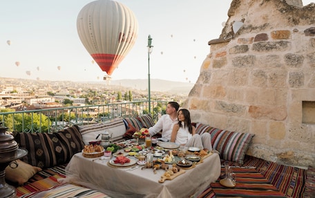 A couple dining on a balcony with a table set for a meal, overlooking a cityscape and a hot air balloon in the distance.