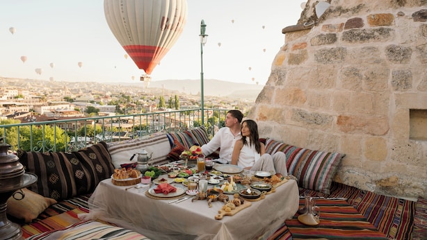 Een stel eet aan een balkon met een gedekte tafel, met uitzicht op een stadsbeeld en een luchtballon in de verte.