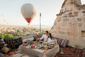 Un couple est assis à une table dressée sur un balcon, prêt à dîner, avec une vue sur la ville et un ballon à air chaud qui flotte au loin.