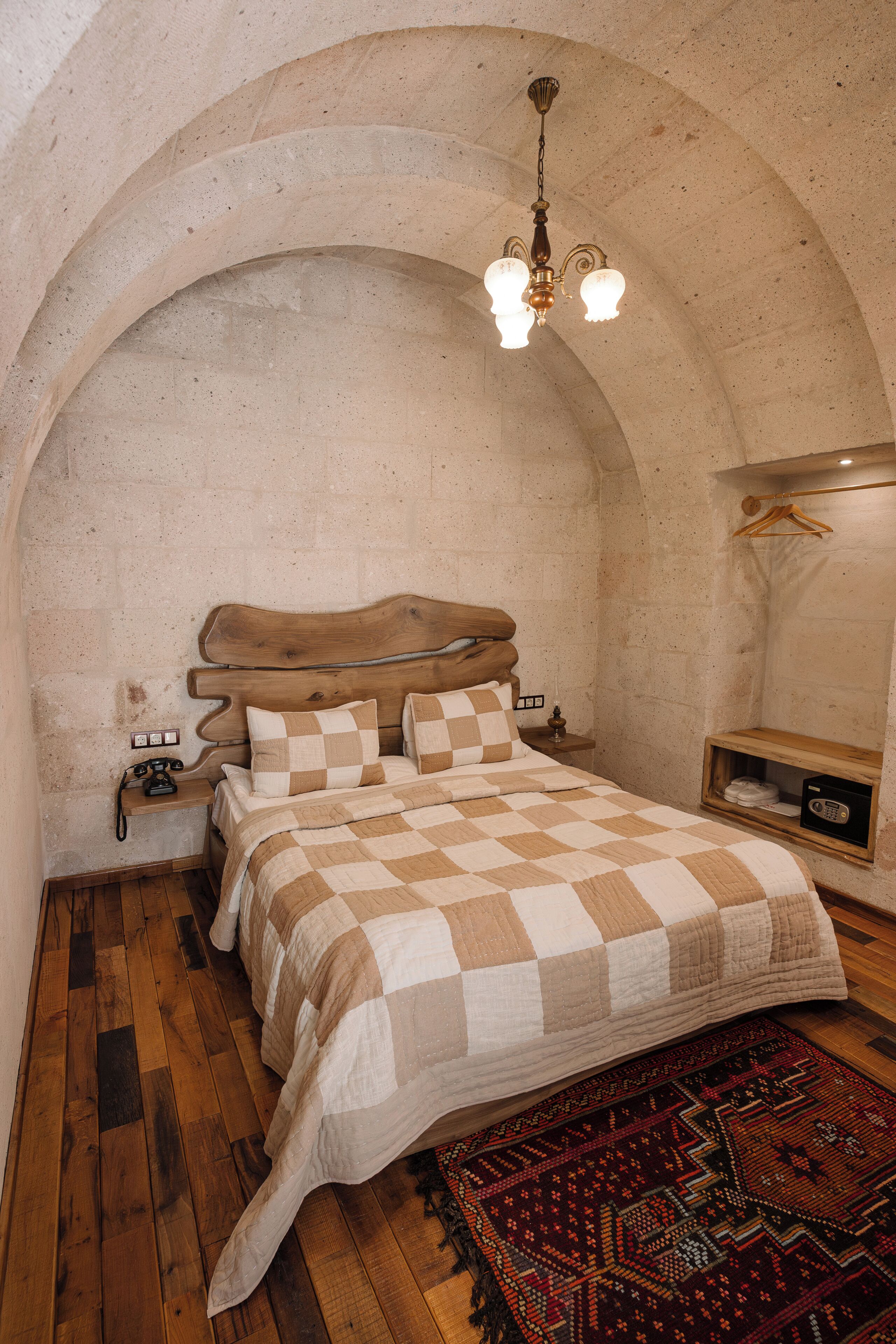 Un dormitorio con colcha a cuadros, cabecero de madera, lámpara colgante, mesita de noche de madera y alfombra estampada.