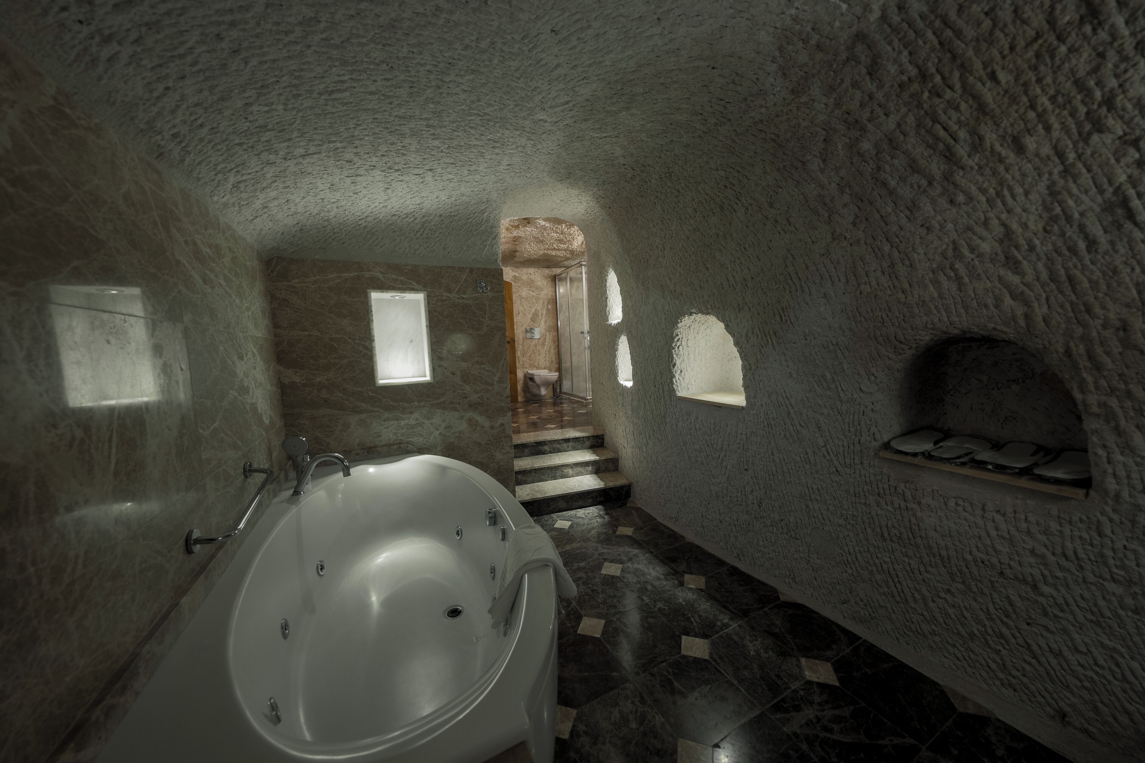 A bathroom with a large bathtub, a shower, and a stone wall. There is a small window and a shelf with stones.