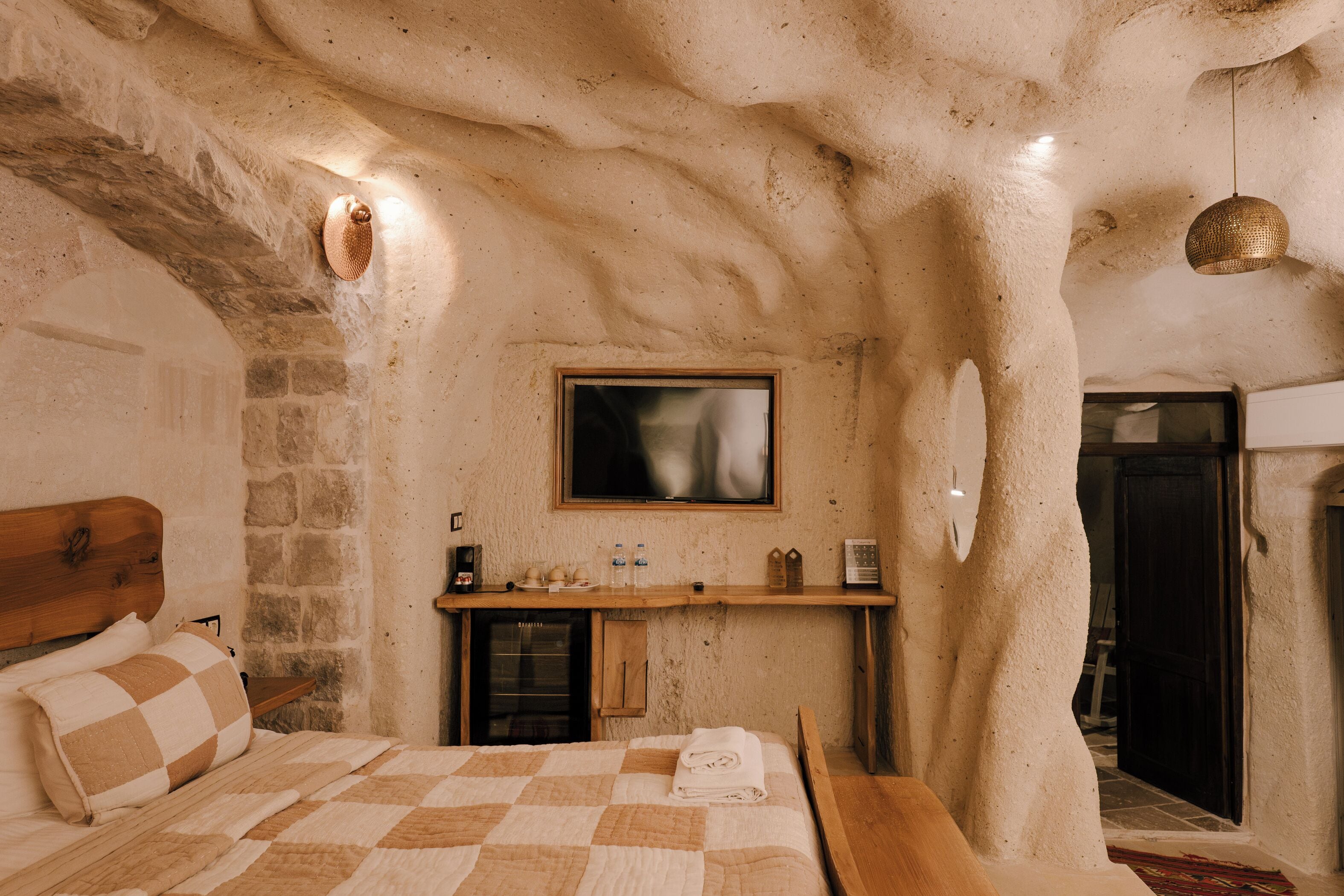 A bedroom with a stone wall, a bed with a checkered quilt, a wooden headboard, a small shelf with toiletries, and a television mounted on the wall.