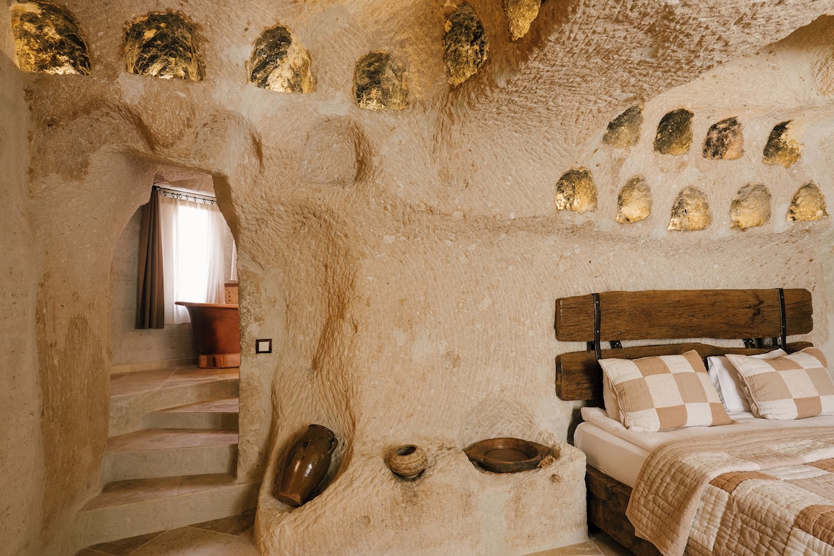 Una habitación rústica con pared de piedra, una cama de madera con almohadones de cuadros, un banco de piedra y una ventana pequeña con cortinas.
