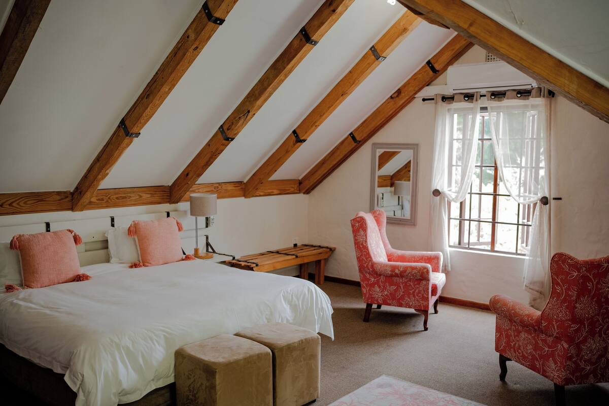 Un dormitorio con una cama grande, dos sillas, un banco de madera y una ventana con cortinas.