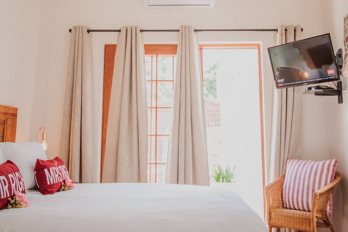Un dormitorio con cama, televisor montado en la pared, ventana con cortinas y techo de madera.