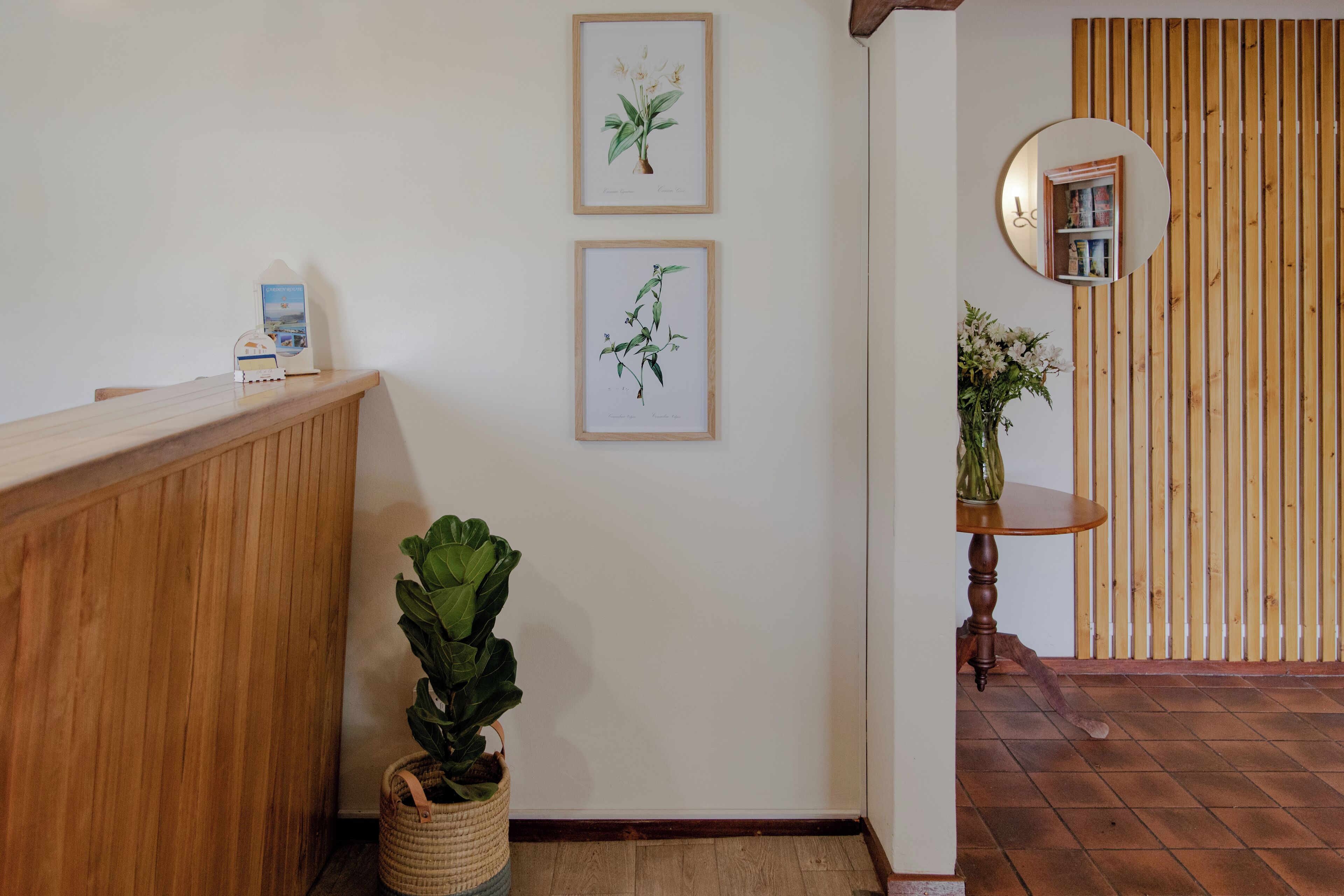 Zona de recepción con mostrador de madera, dos ilustraciones botánicas enmarcadas, una planta en maceta, un espejo redondo y un revestimiento de pared de listones de madera.