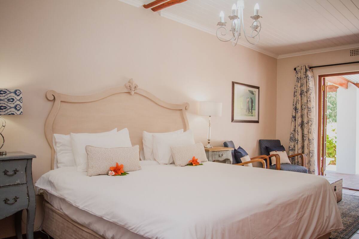 Una cama bien hecha con sábanas blancas y flores naranjas, una mesita de noche de madera con una lámpara, una silla y un cuadro en la pared.