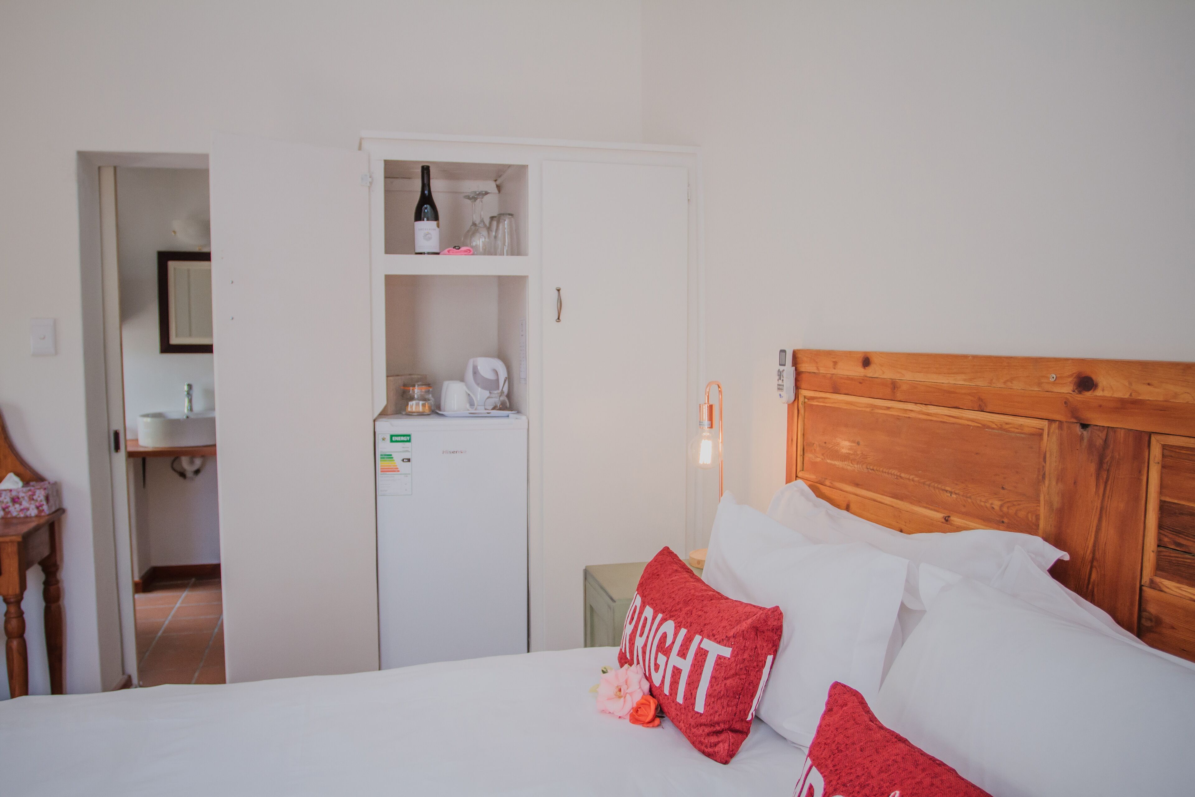 A hotel room with a wooden headboard, white bedding, a red pillow with the word "BRIGHT," and a minibar with various items.