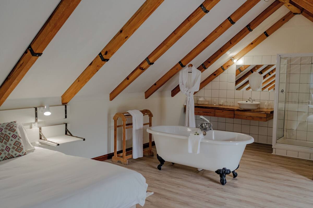 Un dormitorio con techo inclinado, una cama blanca, cabecera de madera, una bañera blanca, un estante de madera y un baño con lavamanos y espejo.