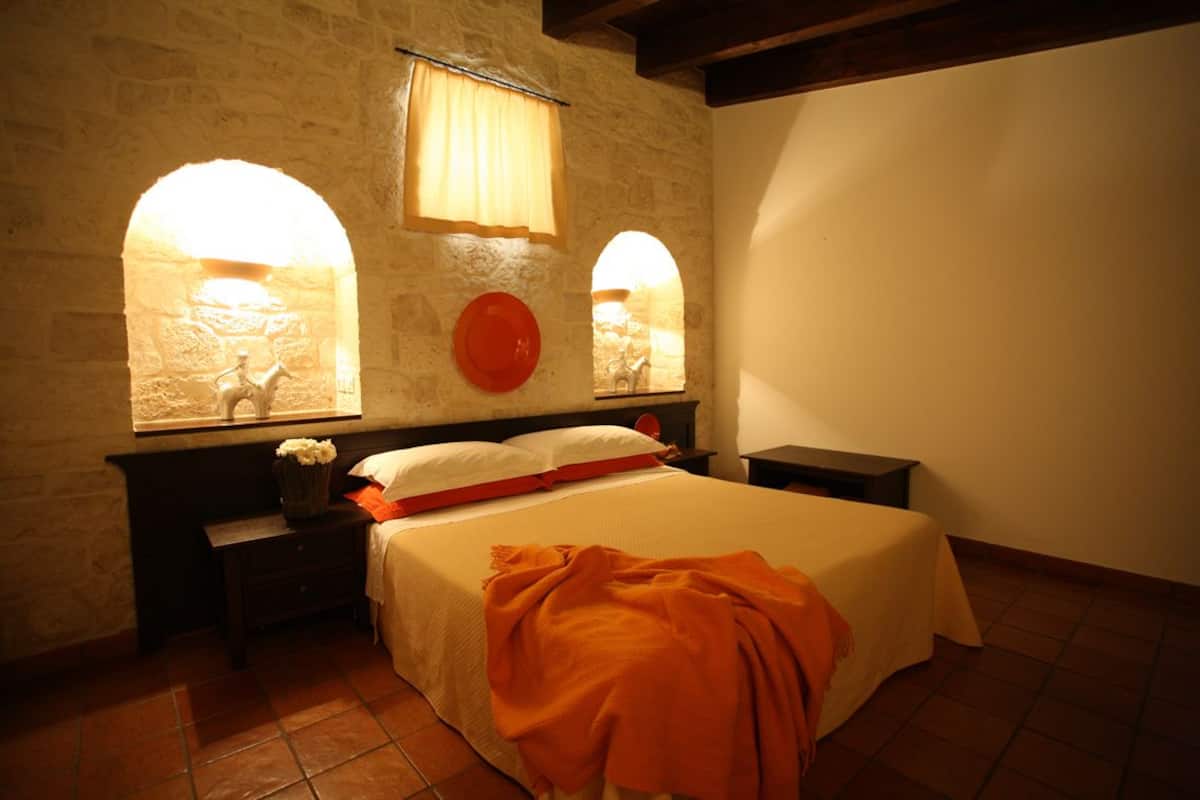 Ein Schlafzimmer mit Steinwand, zwei Nachttischen, einem Bett mit weißer und orangefarbener Bettdecke und zwei Bogenfenstern mit Vorhängen.