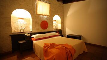 A bedroom with a stone wall, two nightstands, a bed with a white and orange bedspread, and two arched windows with curtains.
