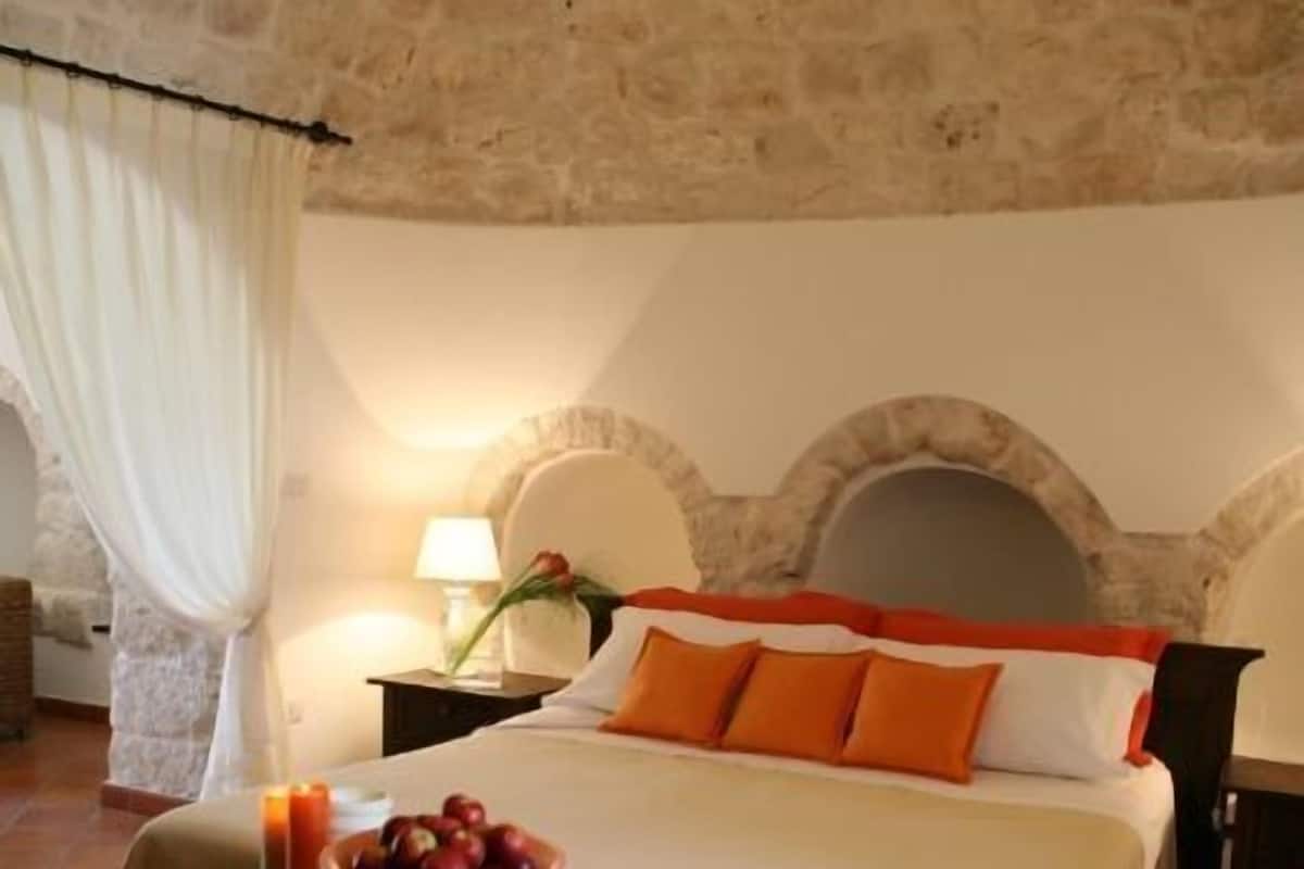 Ein gemütliches Schlafzimmer mit einer Steinsäule, einem Bett mit orange-weißer Bettwäsche, einem Nachttisch mit Kerze und Obst sowie einem Beistelltisch mit Lampe.