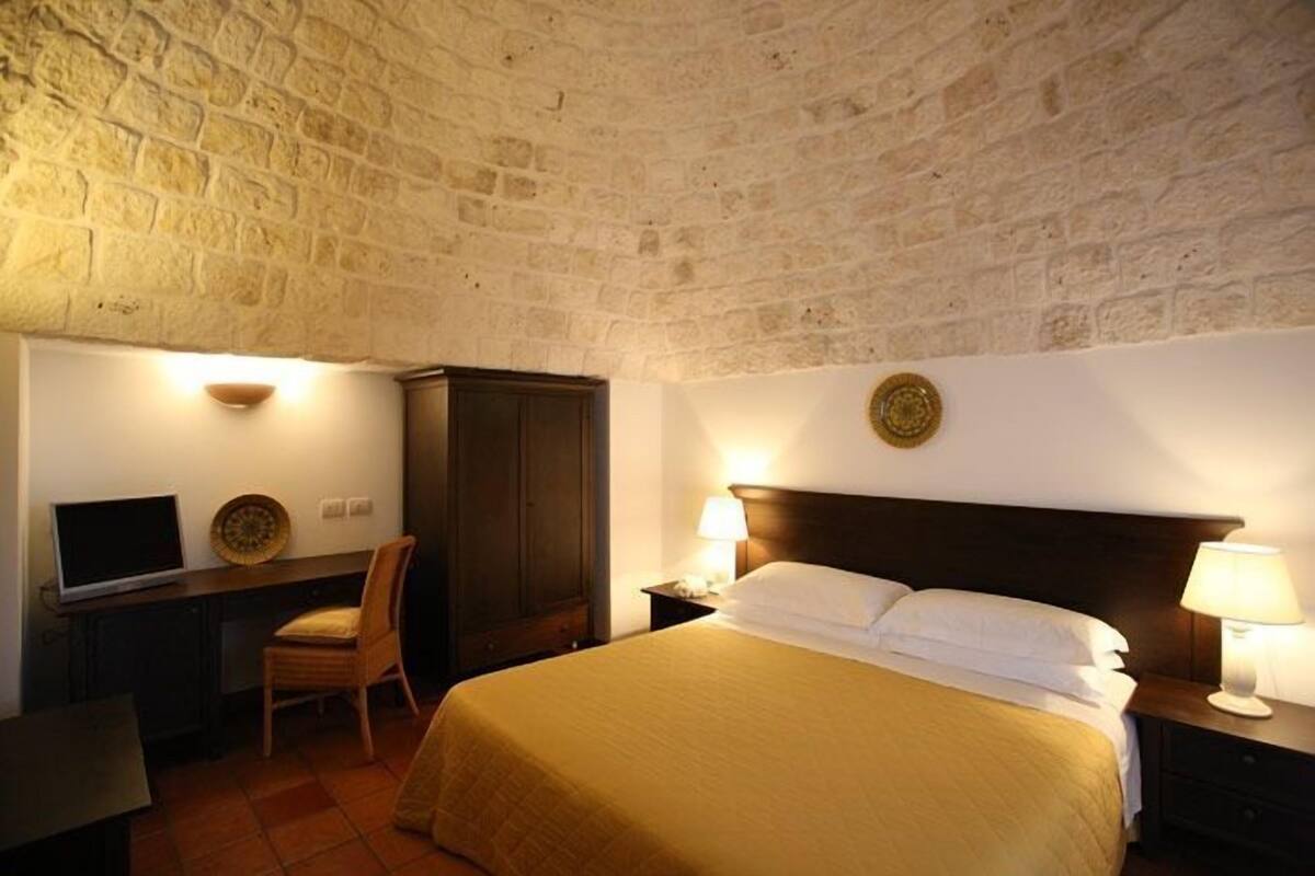 Ein Hotelzimmer mit Steinwand, einem Bett mit weißer Bettwäsche, einem Schreibtisch mit Computer und einem Stuhl.