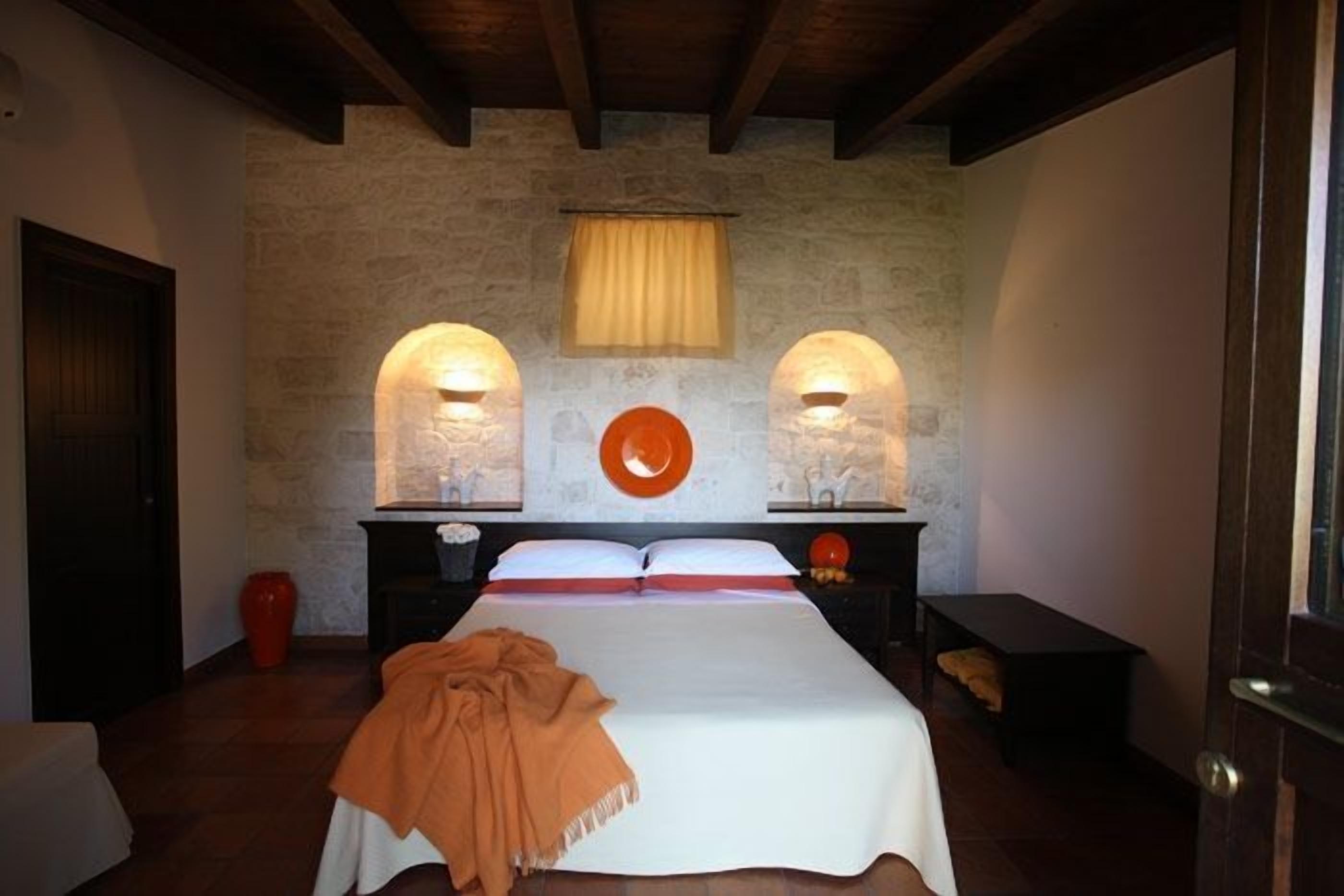 Ein Schlafzimmer mit einer Steinsandsteinwand, zwei Nachttischlampen, einem Bett mit weißer Bettdecke und einer orangefarbenen Tagesdecke sowie einer roten Vase auf dem Nachttisch.