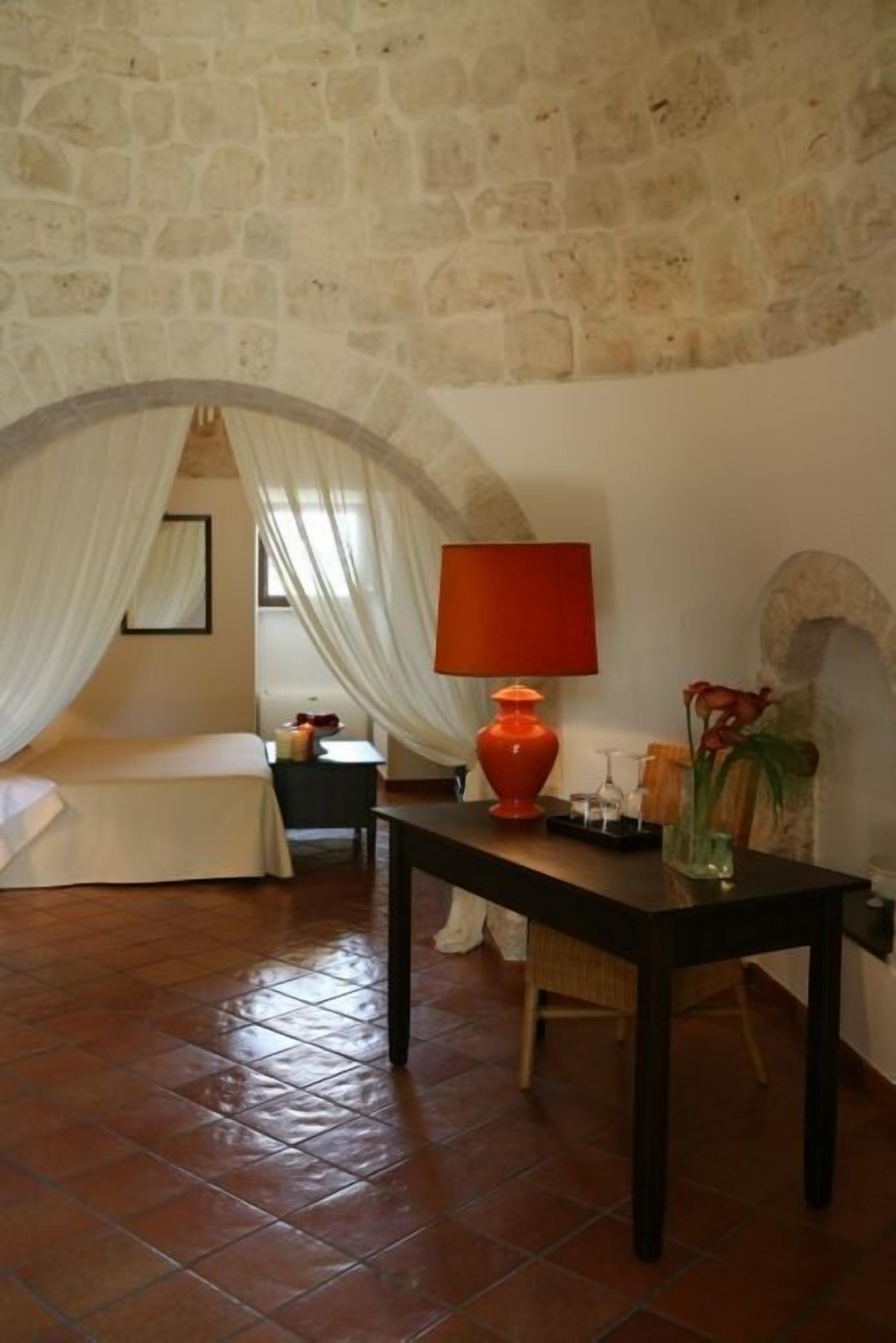Un dormitorio con pared de piedra, cama con ropa blanca, mesa con lámpara roja y un florero con flores.