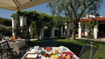 Un área de comedor al aire libre con mesas preparadas para el desayuno, rodeada de follaje y un edificio en el fondo.