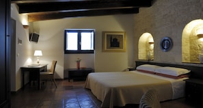 Quarto com cama, escrivaninha, cadeira, janela e televisão presa à parede.