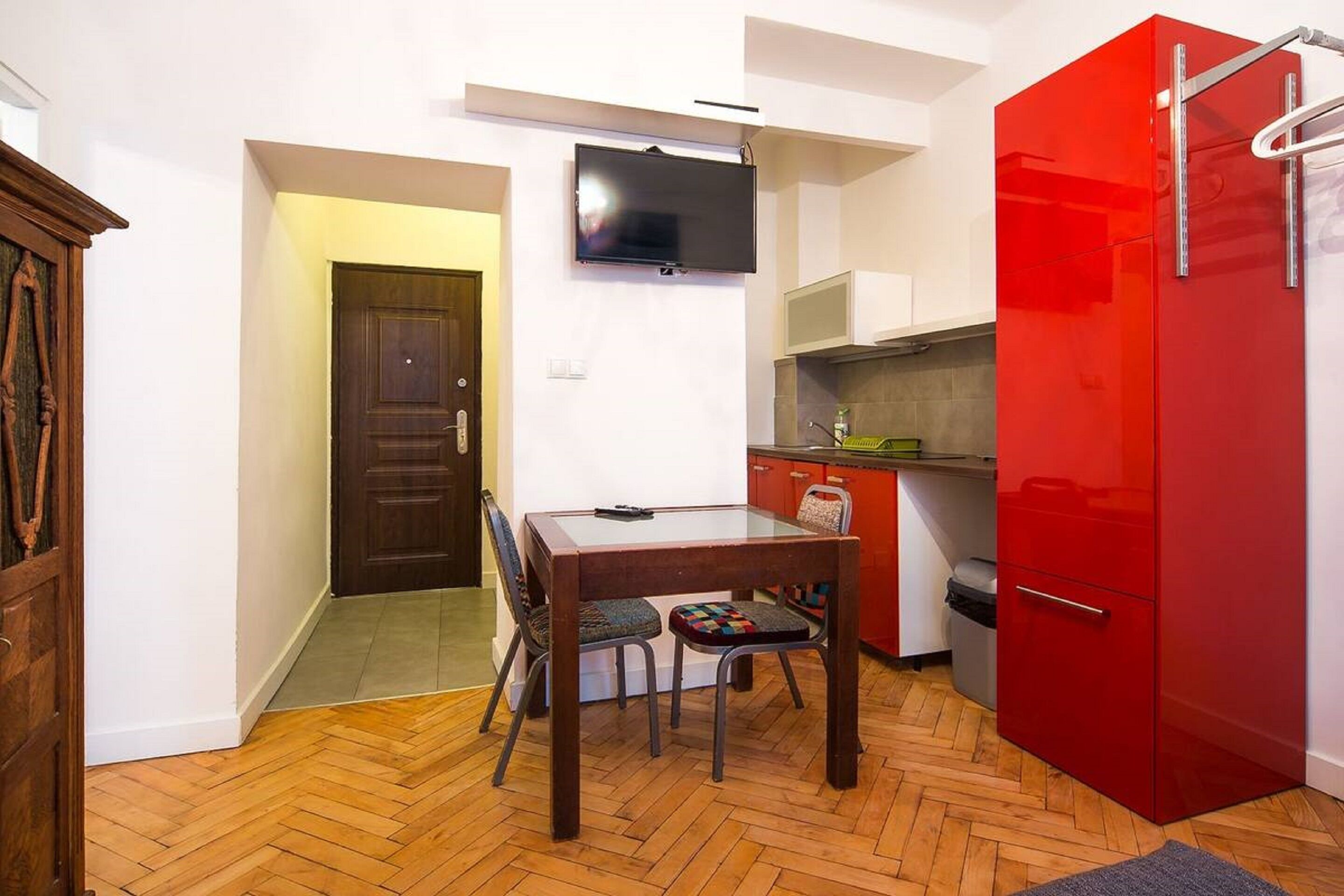 A compact kitchen with a red cabinet, a wooden dining table with two chairs, and a TV mounted on the wall.