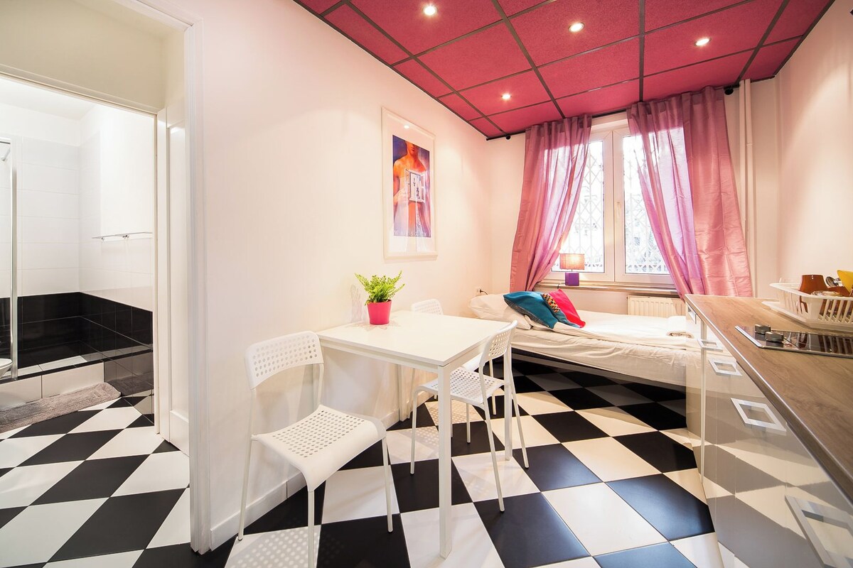 Ein Zimmer mit Schachbrettmuster-Boden, einem Bett mit farbenfrohem Kissen, einem kleinen Tisch mit einer Pflanze und einem Fenster mit rosafarbenen Vorhängen.