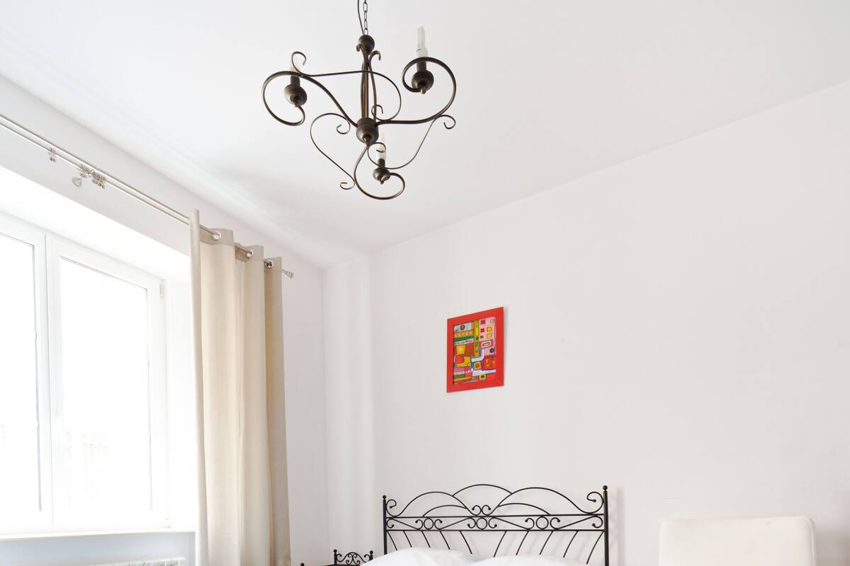 Une chambre à coucher avec un lit, une chaise blanche, un lustre, une fenêtre avec des rideaux et un radiateur.