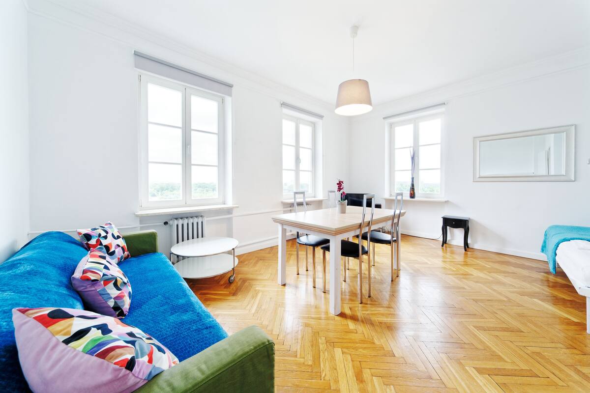 Une pièce lumineuse avec un plancher en bois, un canapé bleu agrémenté de coussins colorés, une table à manger avec des chaises et de grandes fenêtres.