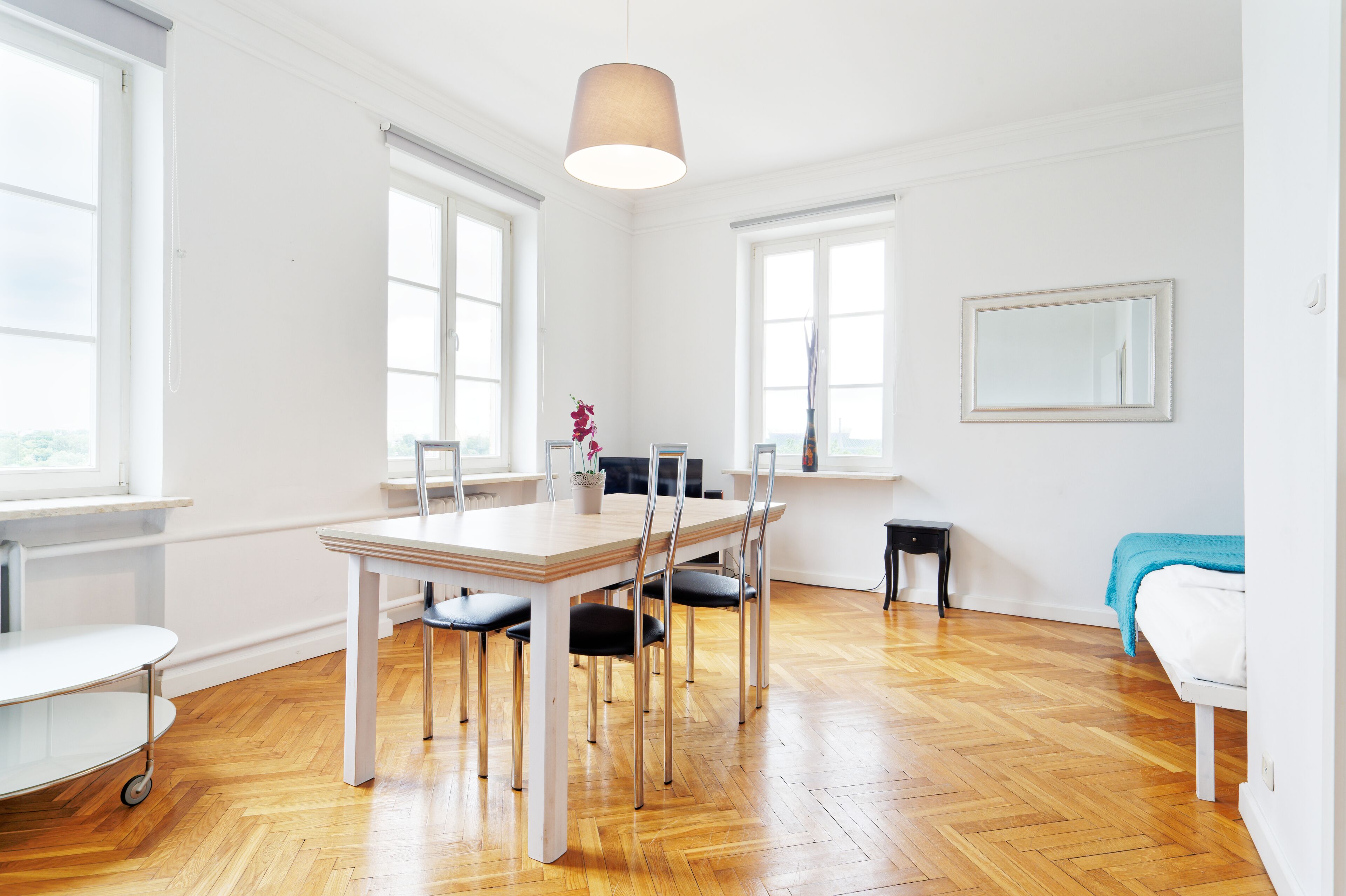 Una habitación luminosa y espaciosa con suelo de madera, una mesa de comedor con sillas, una mesita auxiliar pequeña, una cama con manta azul y ventanas grandes.