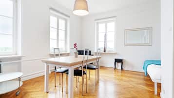 Una habitación luminosa y espaciosa con suelo de madera, una mesa de comedor con sillas, una mesita auxiliar pequeña, una cama con manta azul y ventanas grandes.