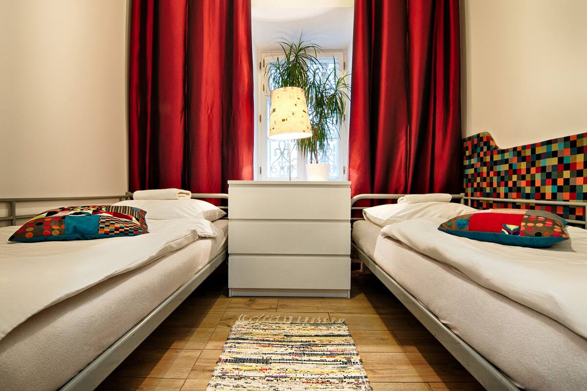 Une chambre avec deux lits, une commode blanche, des rideaux rouges, une plante en pot et un tapis à motifs.