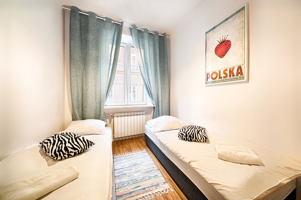Une petite pièce propre, avec deux lits, une fenêtre avec des rideaux, un radiateur et une affiche accrochée au mur.