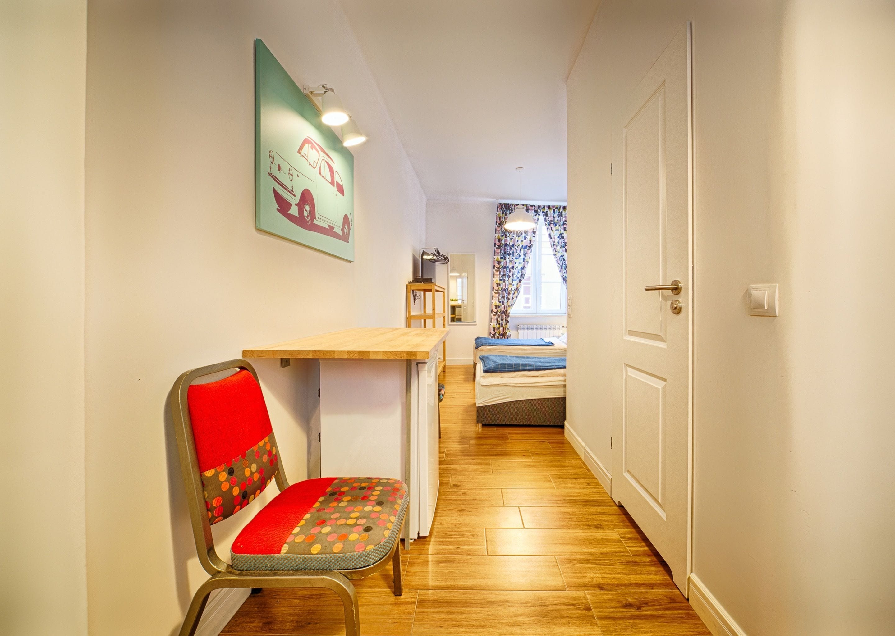 A narrow hallway with a wooden floor, a desk with a chair, a bed with a blue blanket, and a door.
