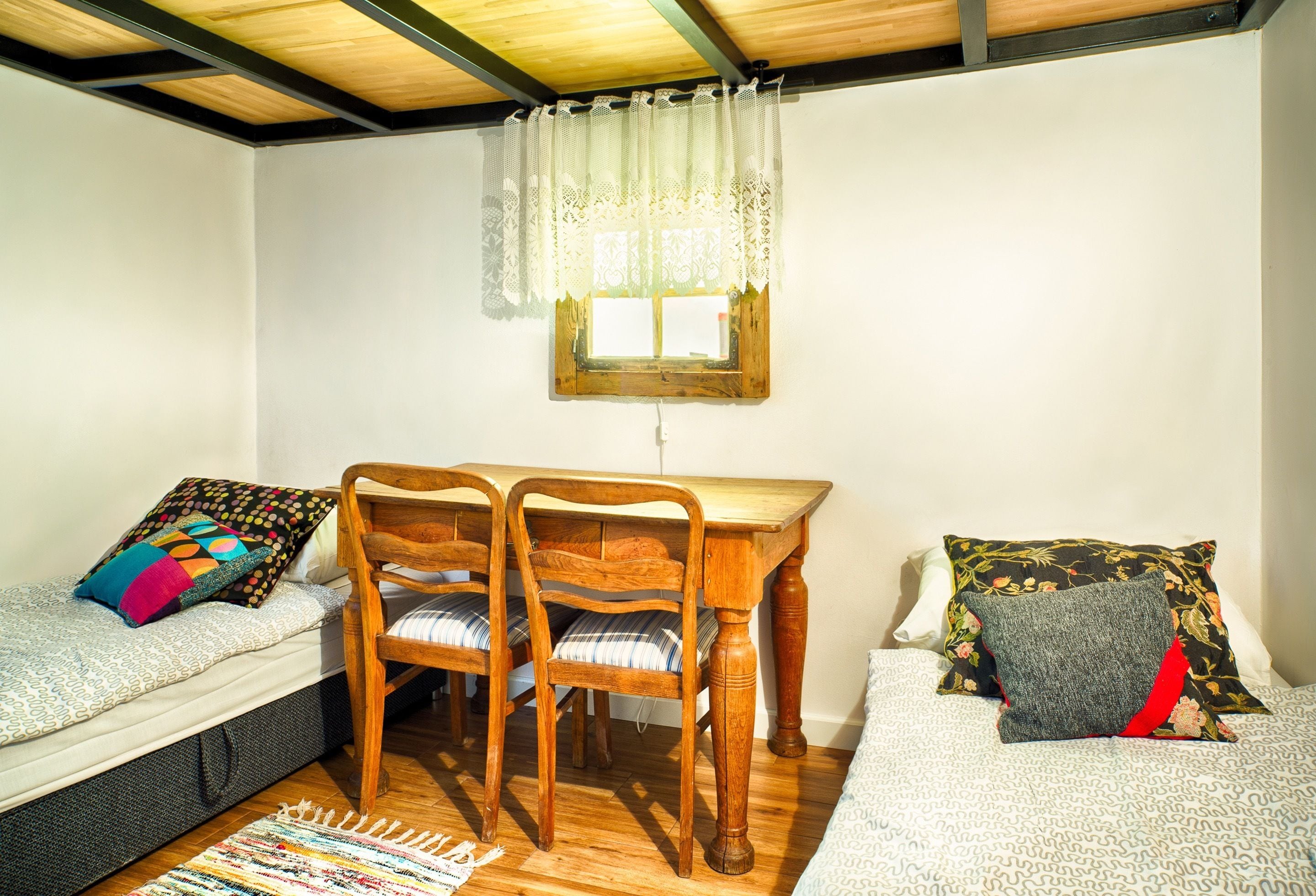 A small, cozy room with a wooden table, two chairs, a bed with patterned bedding, and a window with lace curtains.
