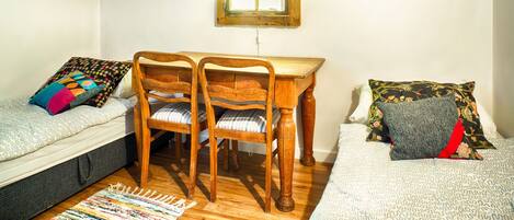 A small, cozy room with a wooden table, two chairs, a bed with patterned bedding, and a window with lace curtains.
