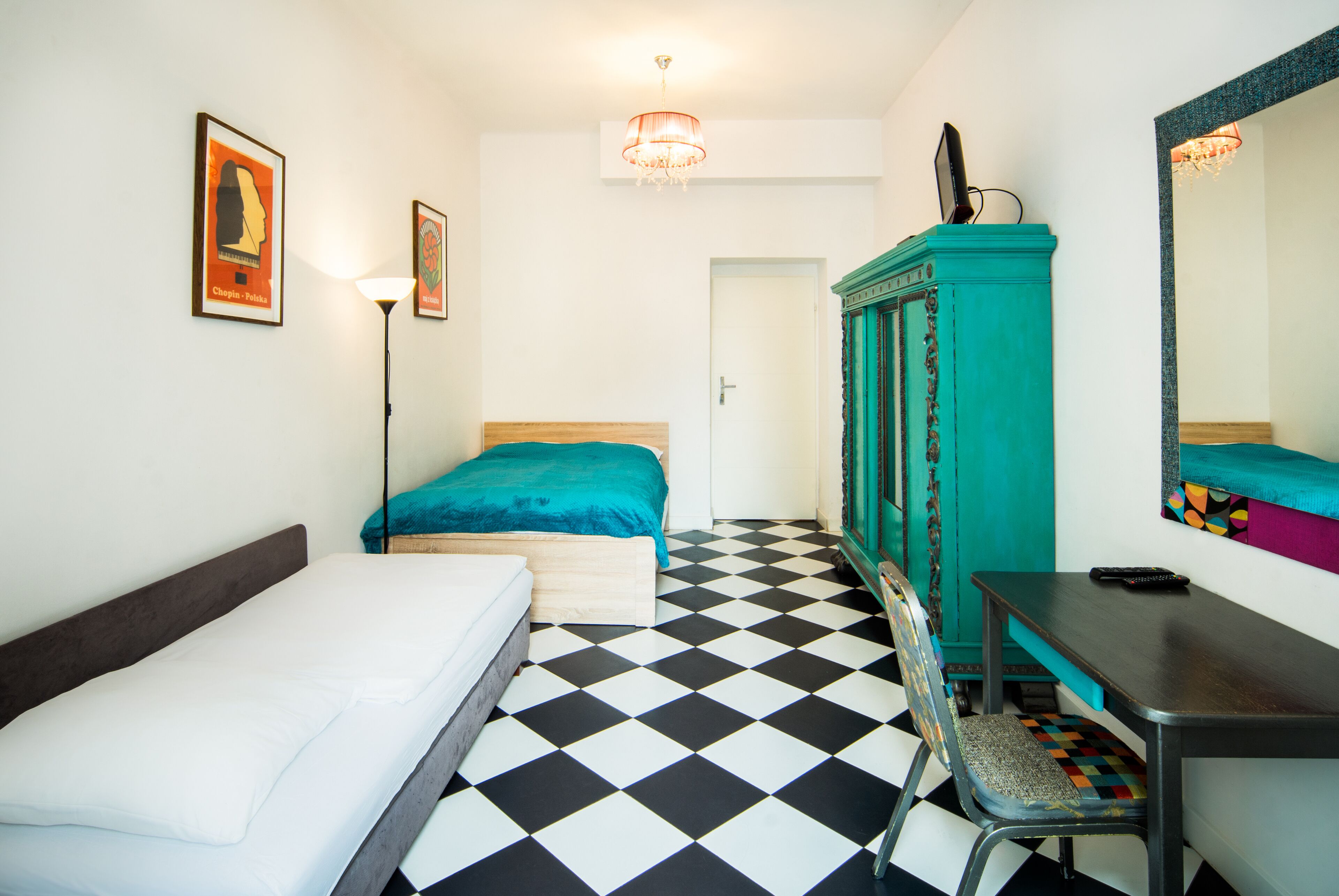A room with a checkered floor, a bed with a teal bedspread, a desk with a chair, a teal wardrobe, and a mirror on the wall.