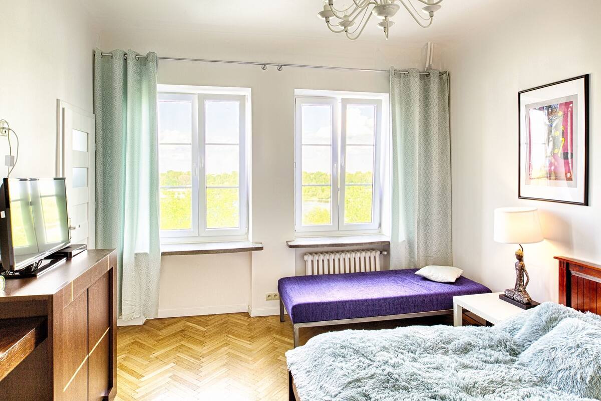 Une pièce avec deux fenêtres, un lit avec un matelas violet, une commode en bois, un téléviseur, un radiateur et un tableau encadré accroché au mur.