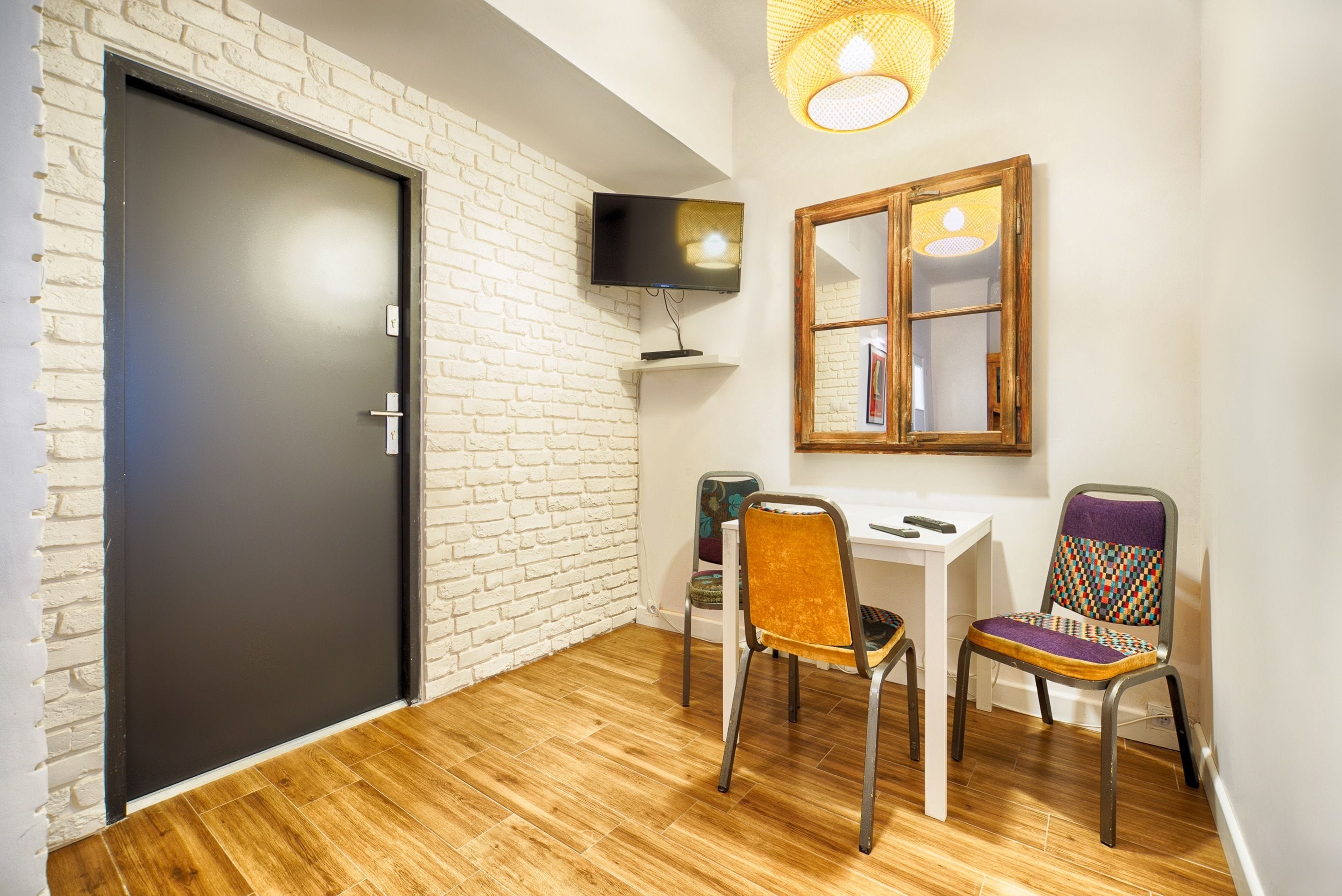 A room with a white brick wall, a dark door, a wooden floor, a white desk, two chairs, a television, and a mirror.