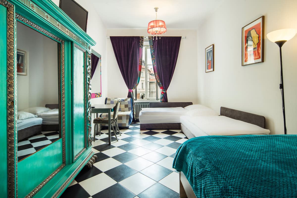 Une chambre d’hôtel avec un sol à carreaux, un lit, une chaise, une table, un miroir et une fenêtre avec des rideaux.