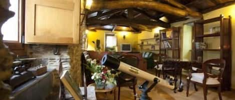 Un interior rústico con chimenea de piedra, vigas de madera, un telescopio y una zona de estar acogedora con una estantería y sillones.