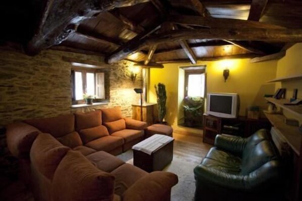 A rustic living room with a stone wall, a brown leather sofa, a green leather armchair, and a television on a wooden stand.