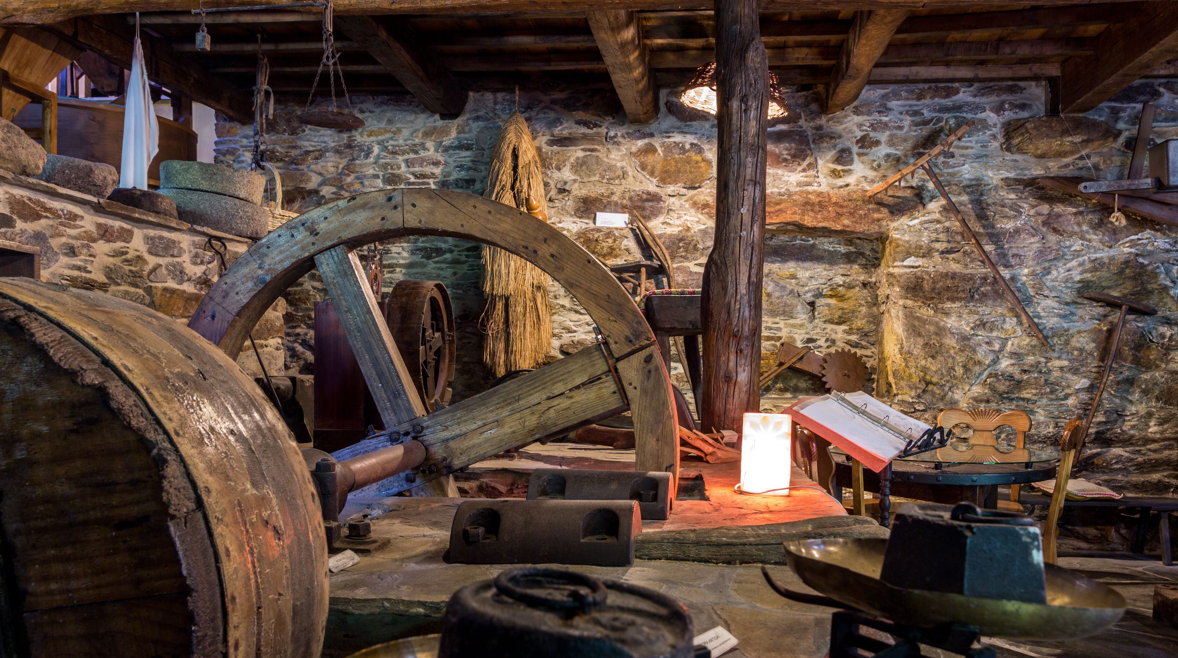 Interno di un edificio storico con pareti in pietra, travi in legno e una grande ruota idraulica.