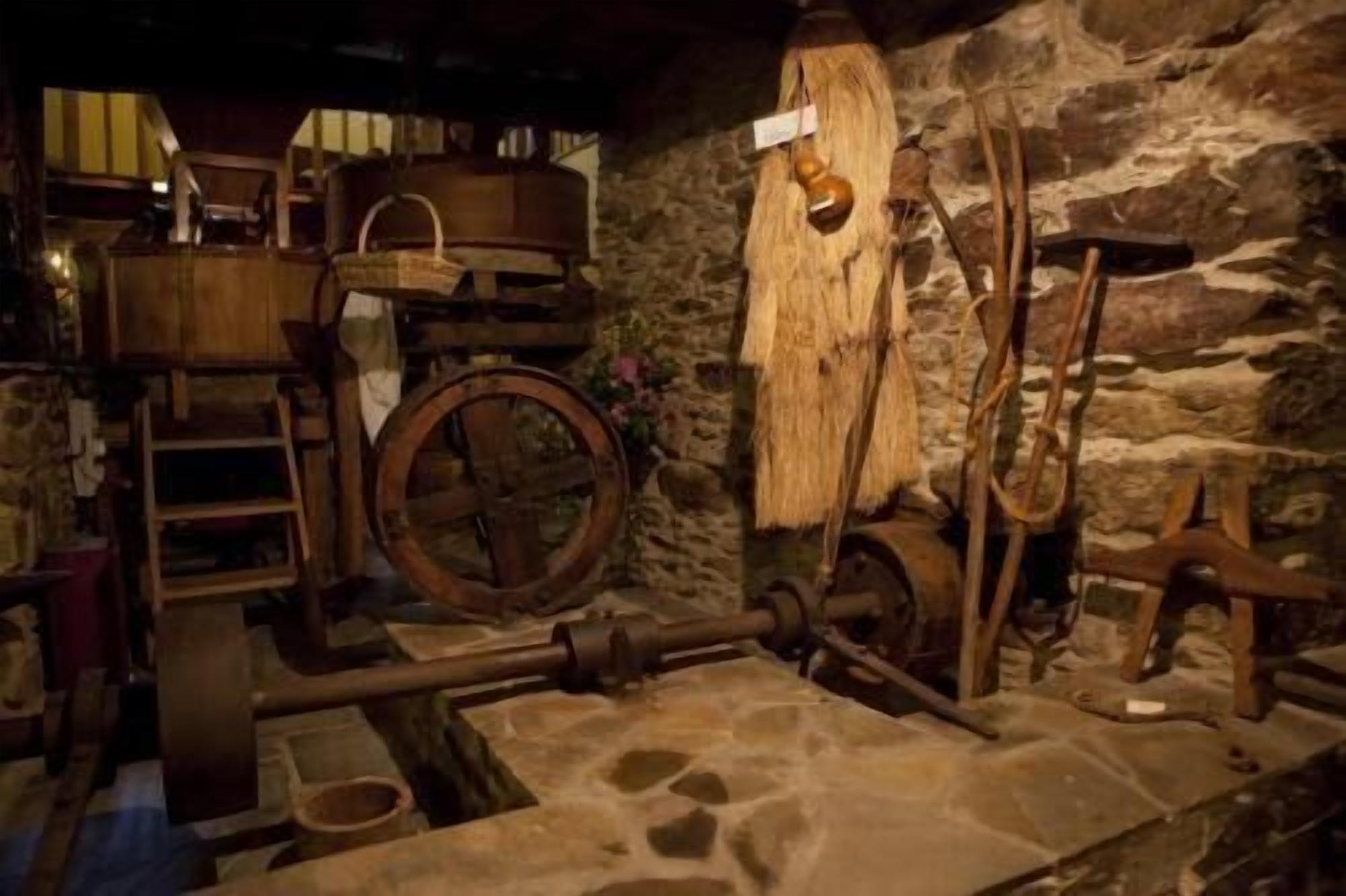 Un interior rústico de piedra con un barril de madera, una rueda grande y una estructura de madera que recuerda a un telar.