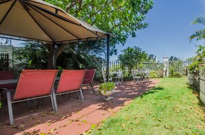 A covered outdoor seating area with red chairs and a table, surrounded by greenery and a clear sky. - HoneyPot Guesthouse (Umhlanga)