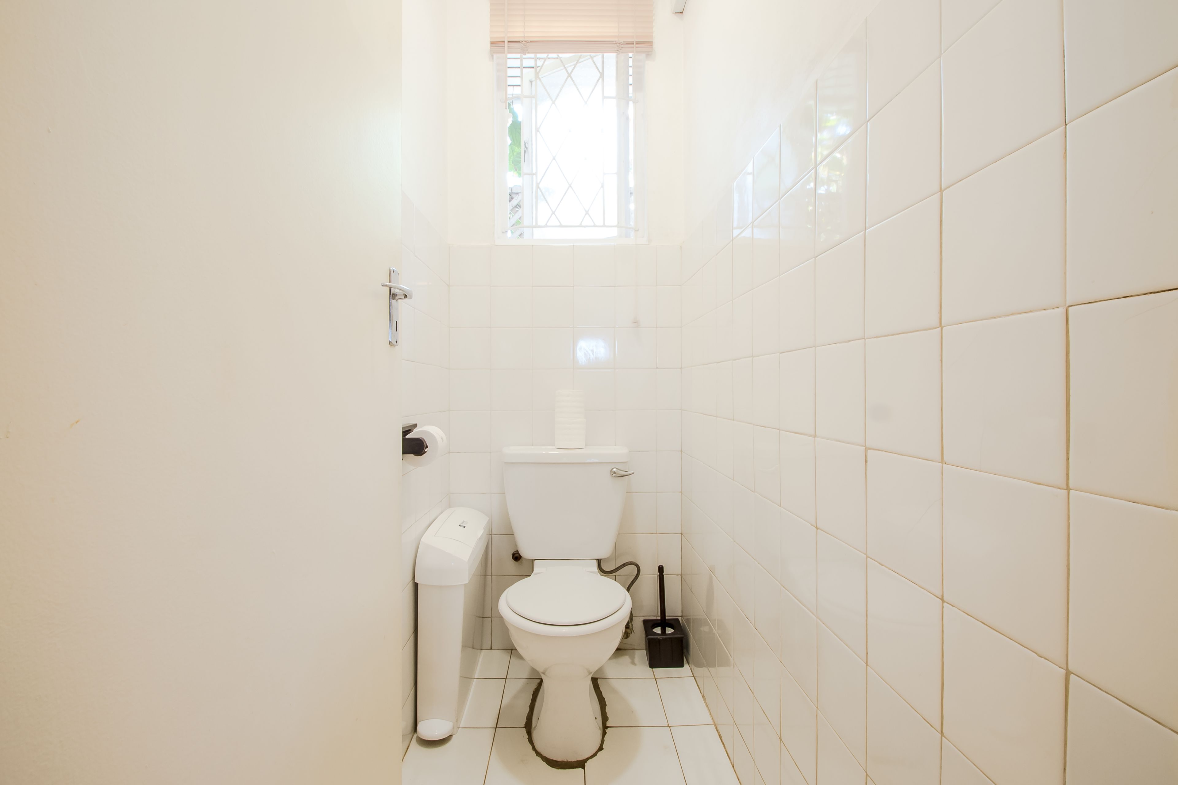 A small, clean bathroom with white tiles, a toilet, a trash bin, and a window with a metal grille.