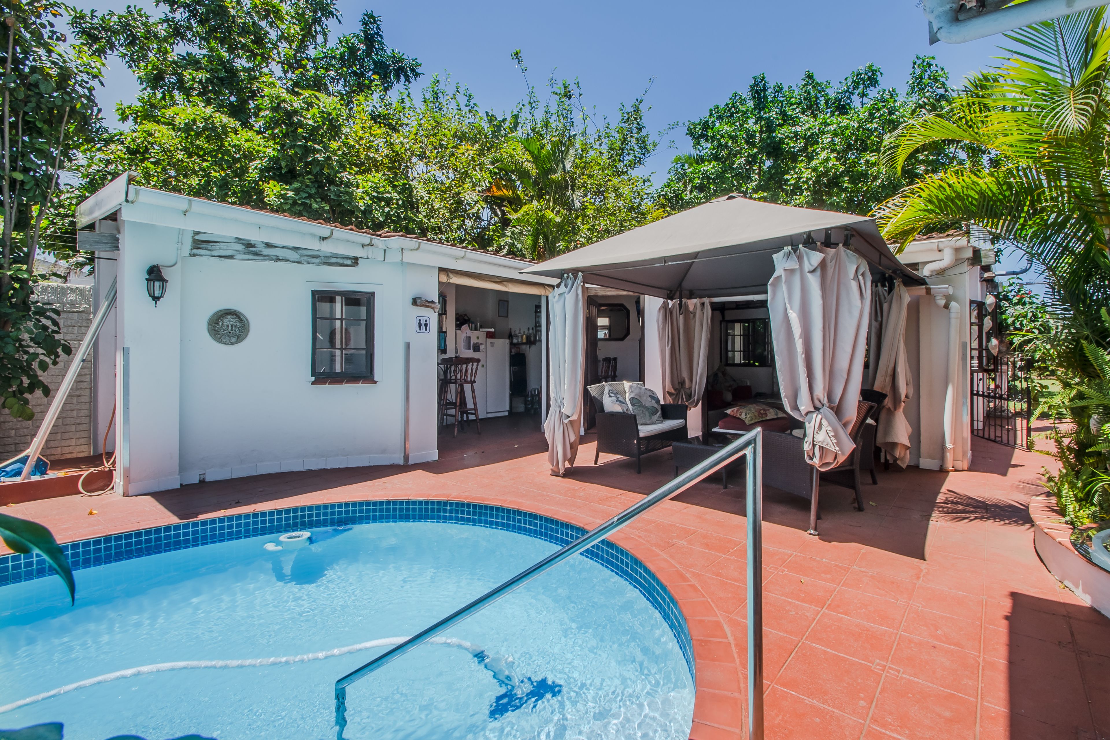 A pool area with a white building, a covered seating area, and a patio with outdoor furniture.