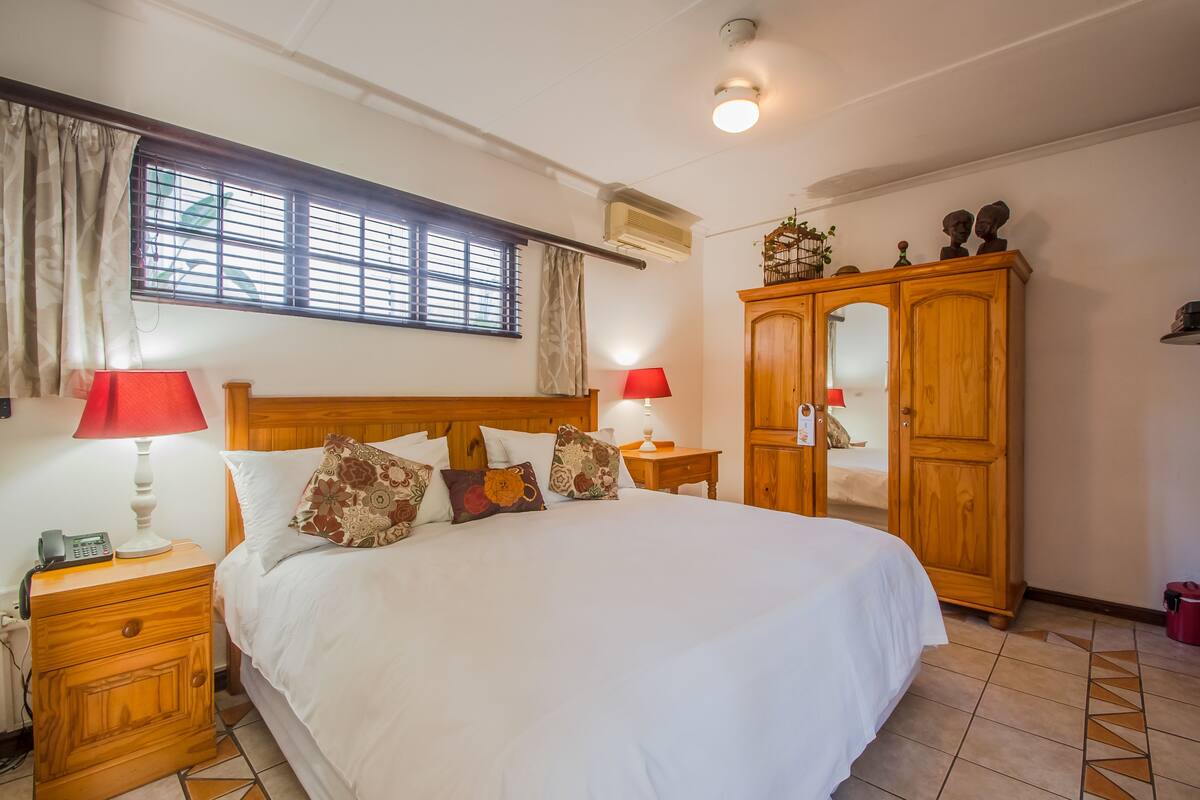A well-lit bedroom with a large bed, two bedside lamps, a wooden wardrobe, and a window with curtains.