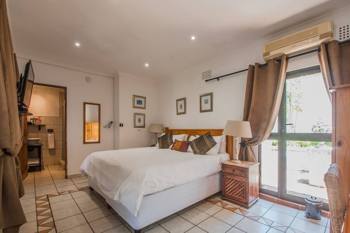 A hotel room with a large bed, a wooden nightstand, a bathroom with a shower, and a view through the window.